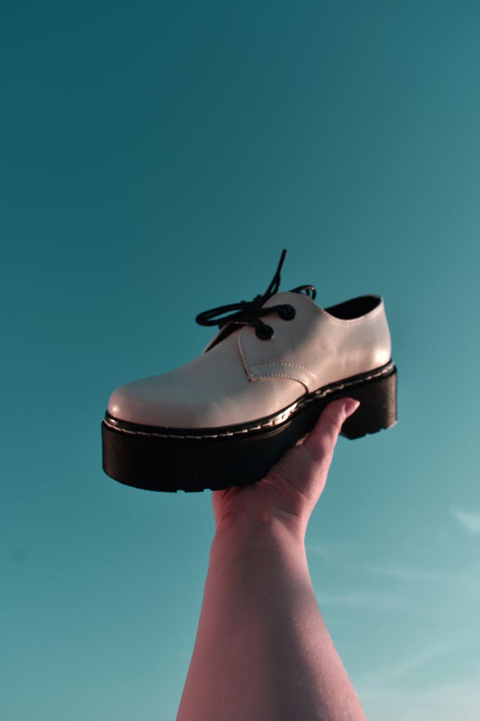 Hand holding a fashionable leather shoe against a vibrant blue sky, emphasizing modern style.